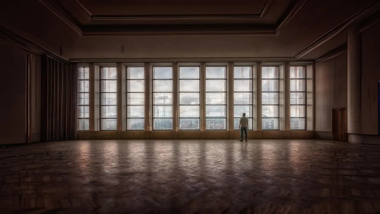 Lone figure at a wall of tall windows across an empty hall with a parquet floor