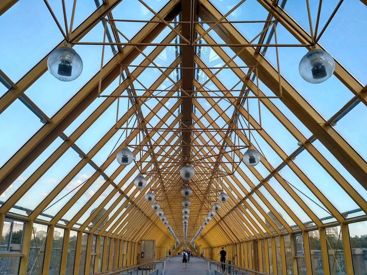 Dugačak prolaz s krovom od stakla pod žutom čeličnom rešetkom, gledano odozdo