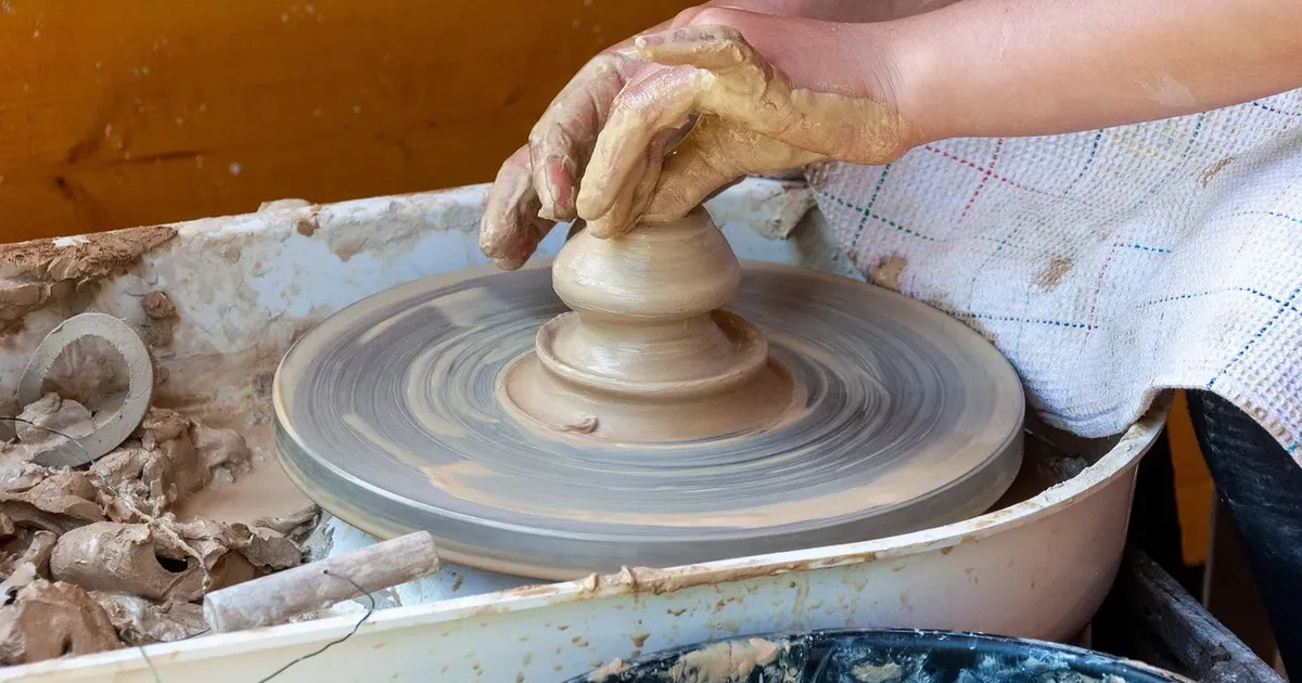 Hands shaping a small clay vessel on a potter's wheel in a clay-spattered basin