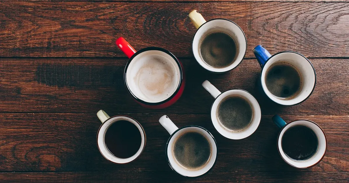 Seven enamel mugs of coffee from above on a dark wooden table