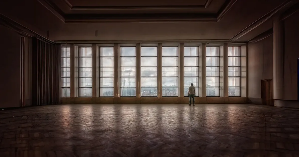 Lone figure at a wall of tall windows across an empty hall with a parquet floor