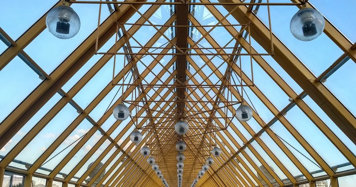 Long glass-roofed walkway under a yellow steel lattice, seen from below